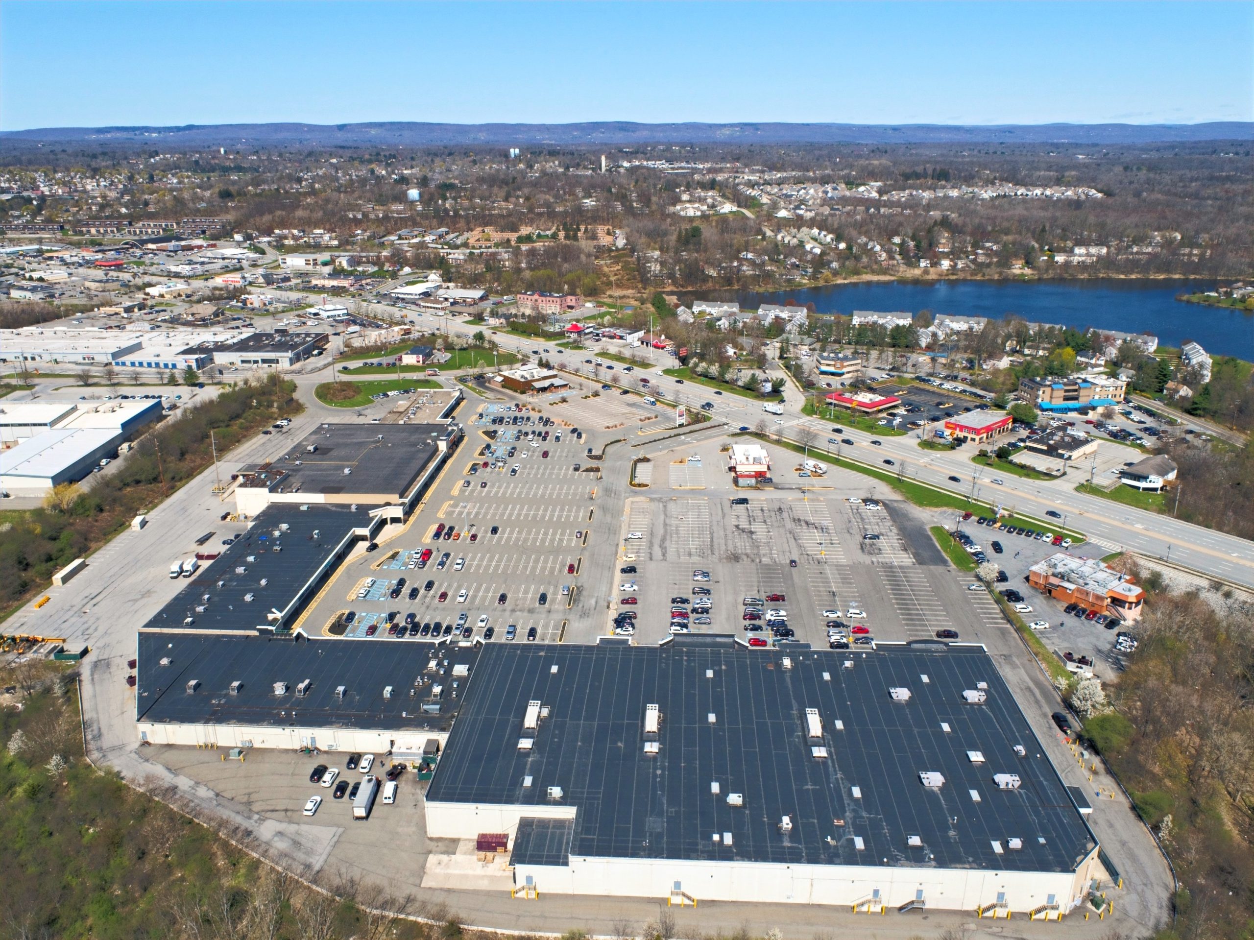 Aerial view of a large shopping complex with parking lots, surrounded by roads, trees, and a distant lake under a clear blue sky.