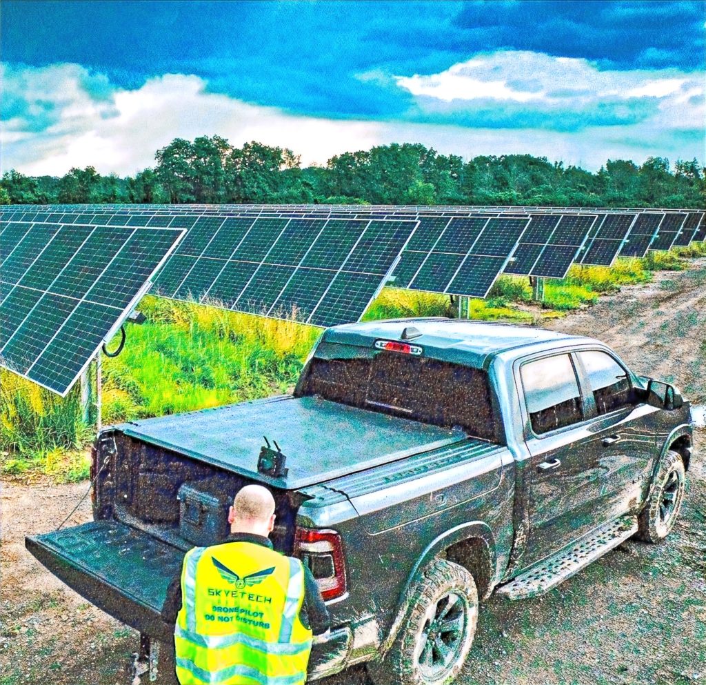 Man in neon vest by truck near solar panels under a cloudy sky. Vest text: "Skytech Drone Pilot Do Not Disturb." Green landscape.