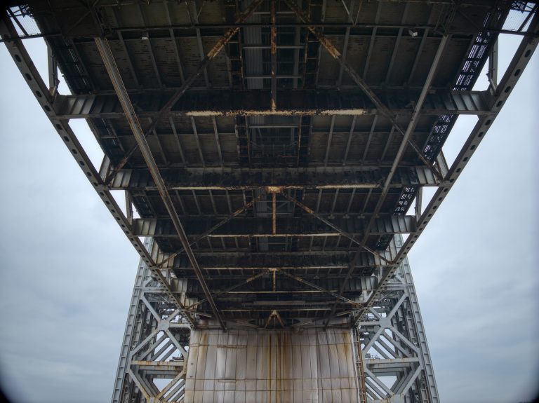 Underneath view of a rusty metal bridge against a cloudy sky. Steel beams and girders create a geometric pattern with an industrial feel.