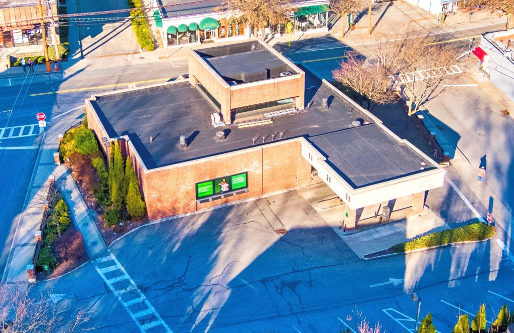 Aerial view of a brick building with a flat roof and green signs. Surrounding streets and bare trees under clear blue skies.