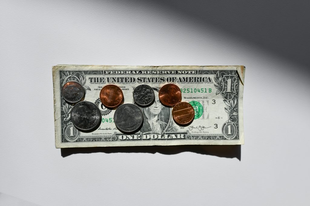 Coins and a dollar bill on a white surface, casting shadows. The bill shows "The United States of America" and "One Dollar."