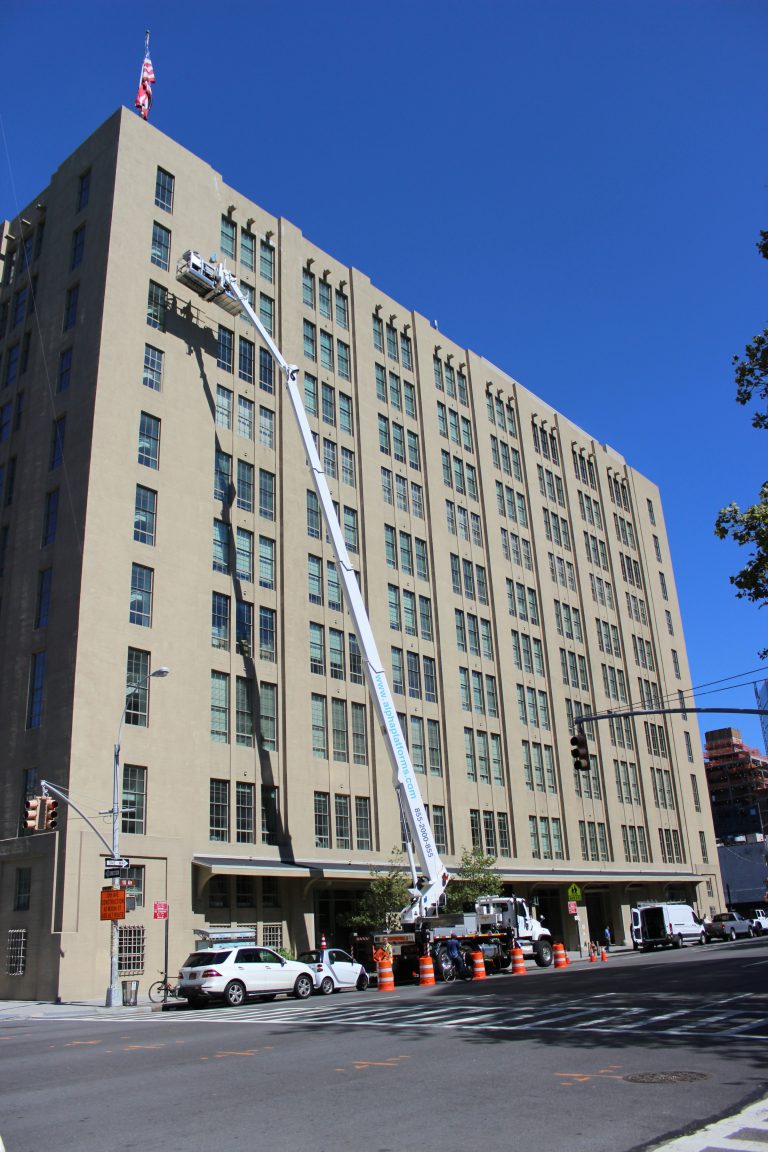 A large beige building with a worker on a lift performing maintenance near the roof; orange cones and utility vehicles are present on the street below.