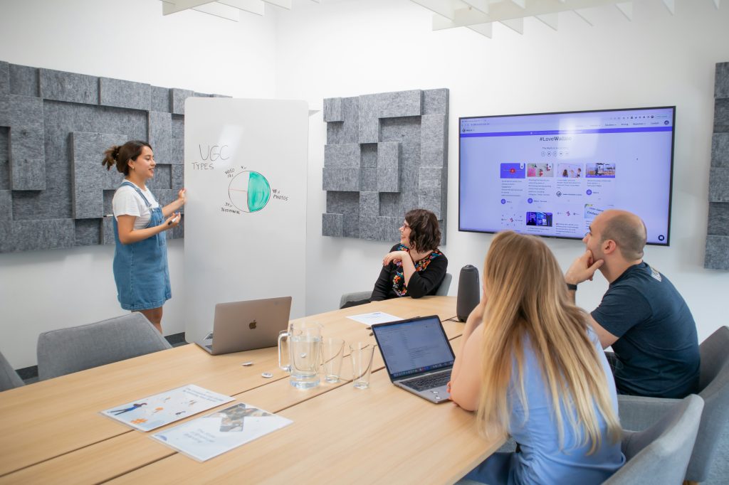 Woman presenting UGC types on whiteboard to three people in a meeting room. Two laptops on the table. Screen displays #ILoveWallis.