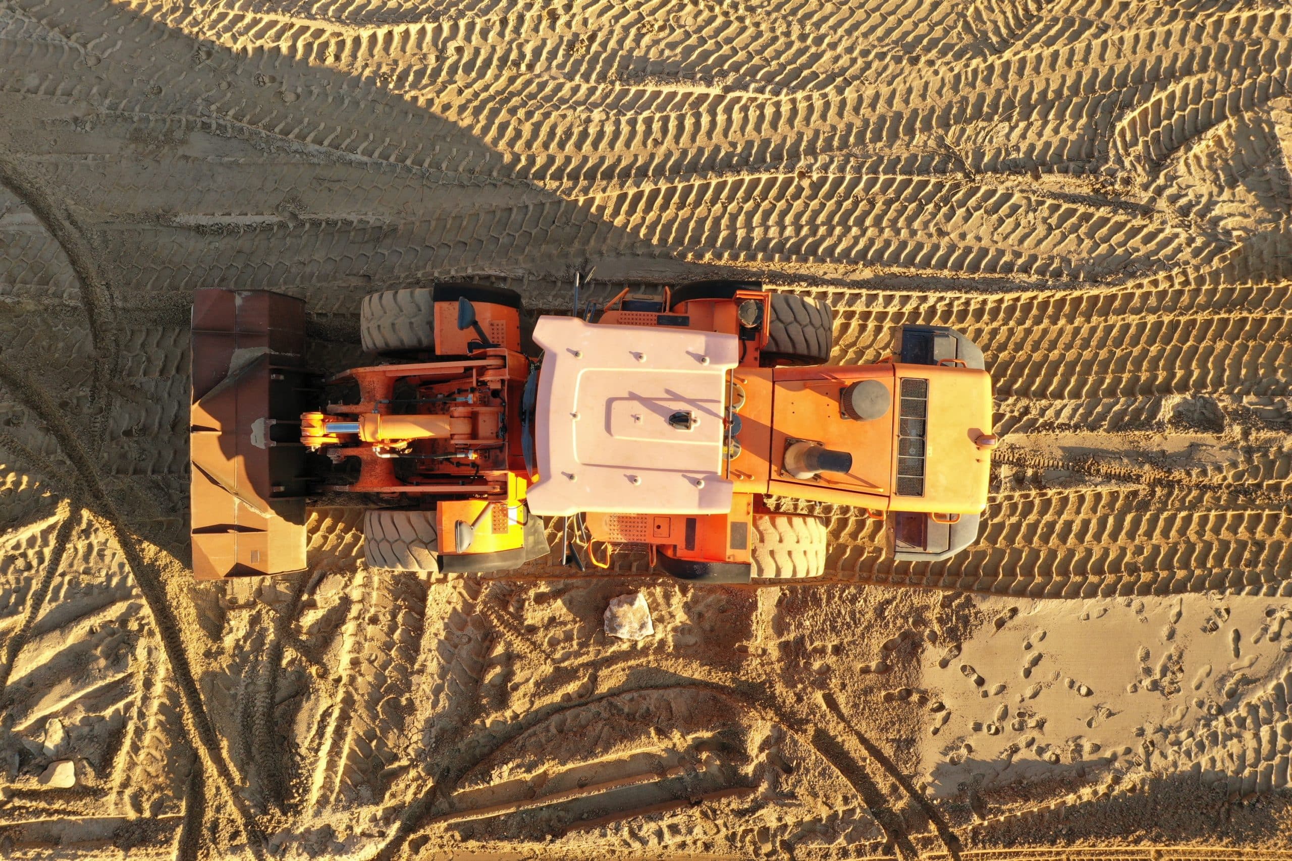 Aerial view of an orange bulldozer on sandy terrain with tire tracks. The sun casts shadows, creating a textured pattern on the ground.