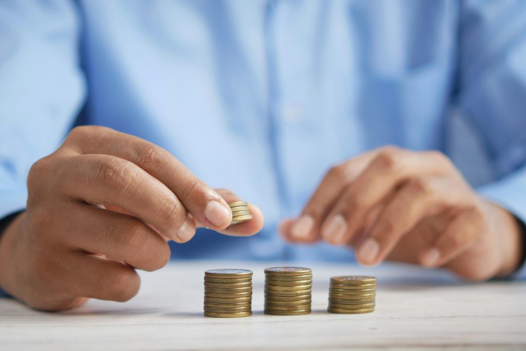 A person in a blue shirt arranges stacks of coins on a white surface, holding several coins in one hand and stacking with the other.