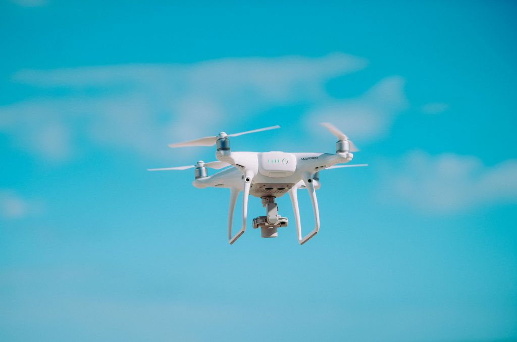 A white quadcopter drone flies in a clear blue sky with soft clouds. Its rotors spin, suggesting movement and technological precision.