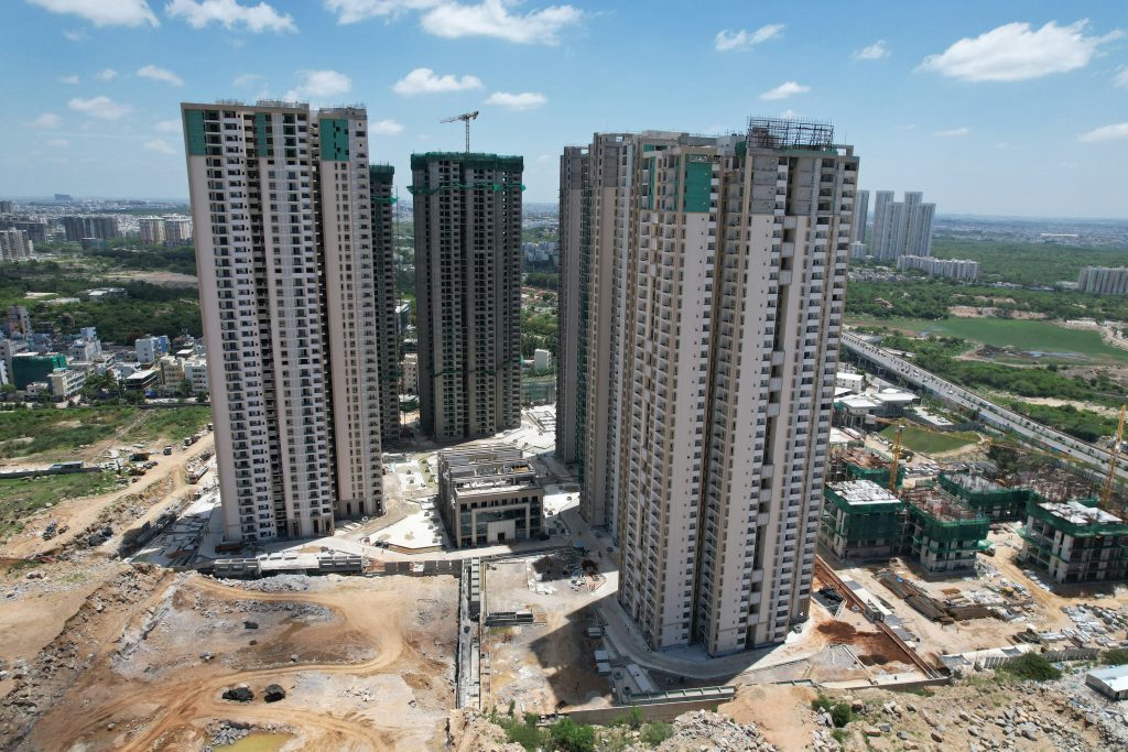 Aerial view of a construction site with several tall, unfinished skyscrapers. The surroundings include green fields and urban areas under a clear blue sky.