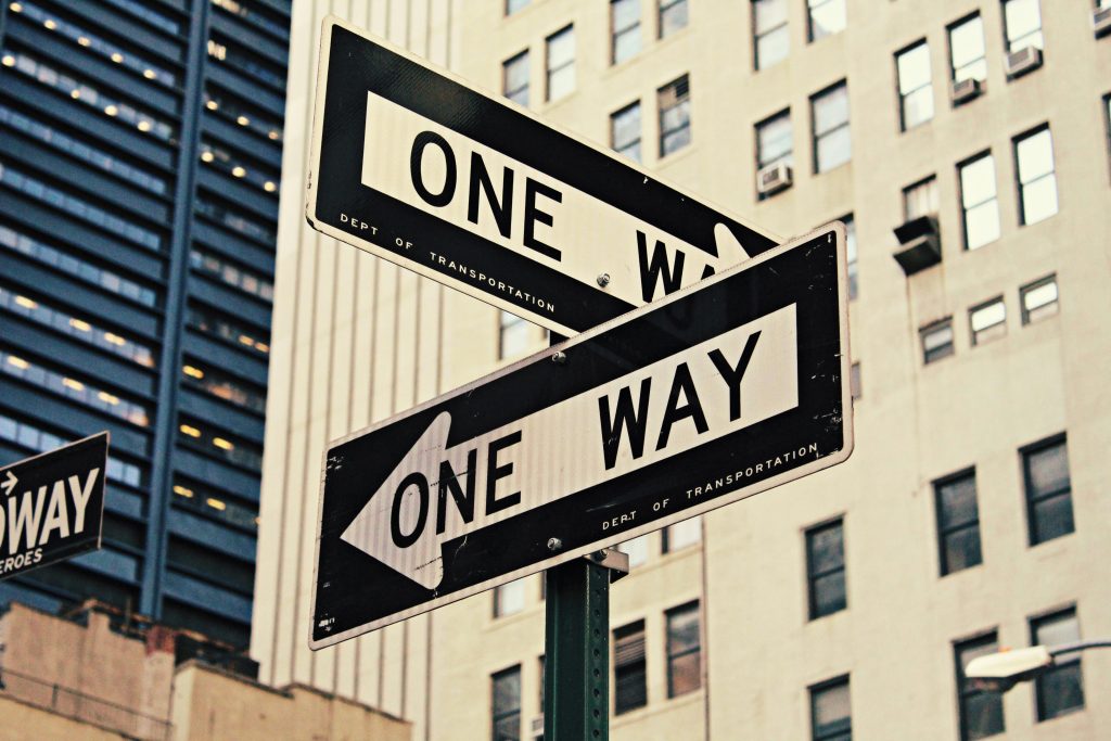 Two intersecting one-way signs point in opposite directions, set against a backdrop of tall urban buildings, conveying a sense of urban complexity.