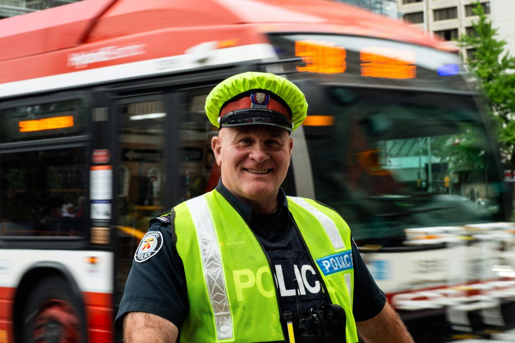 Smiling police officer in a neon vest and cap stands in front of a moving city bus. Urban background with blurred motion, conveying a friendly tone.