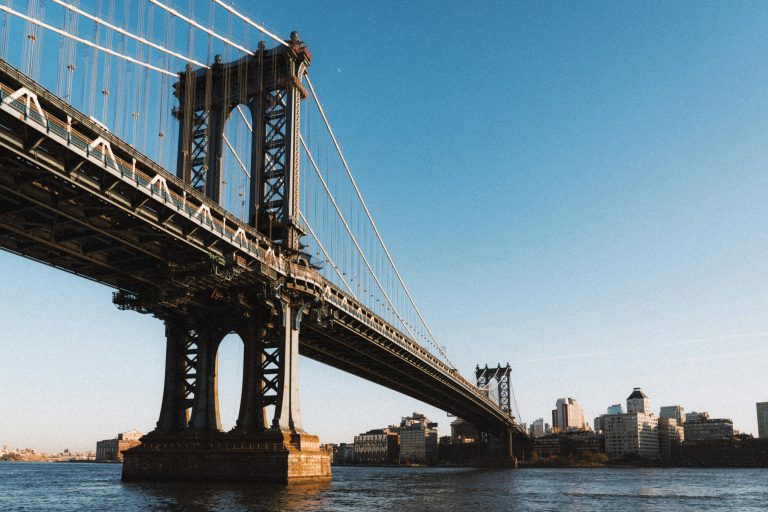 View of a suspension bridge spanning over a river with a city skyline in the background under a clear blue sky, conveying an urban and serene atmosphere.