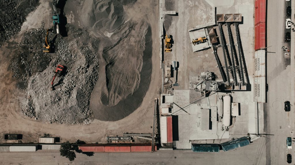 Aerial view of an industrial site with machinery on the left over gravel and dirt. The right side has conveyors and equipment, creating a busy, industrious scene.