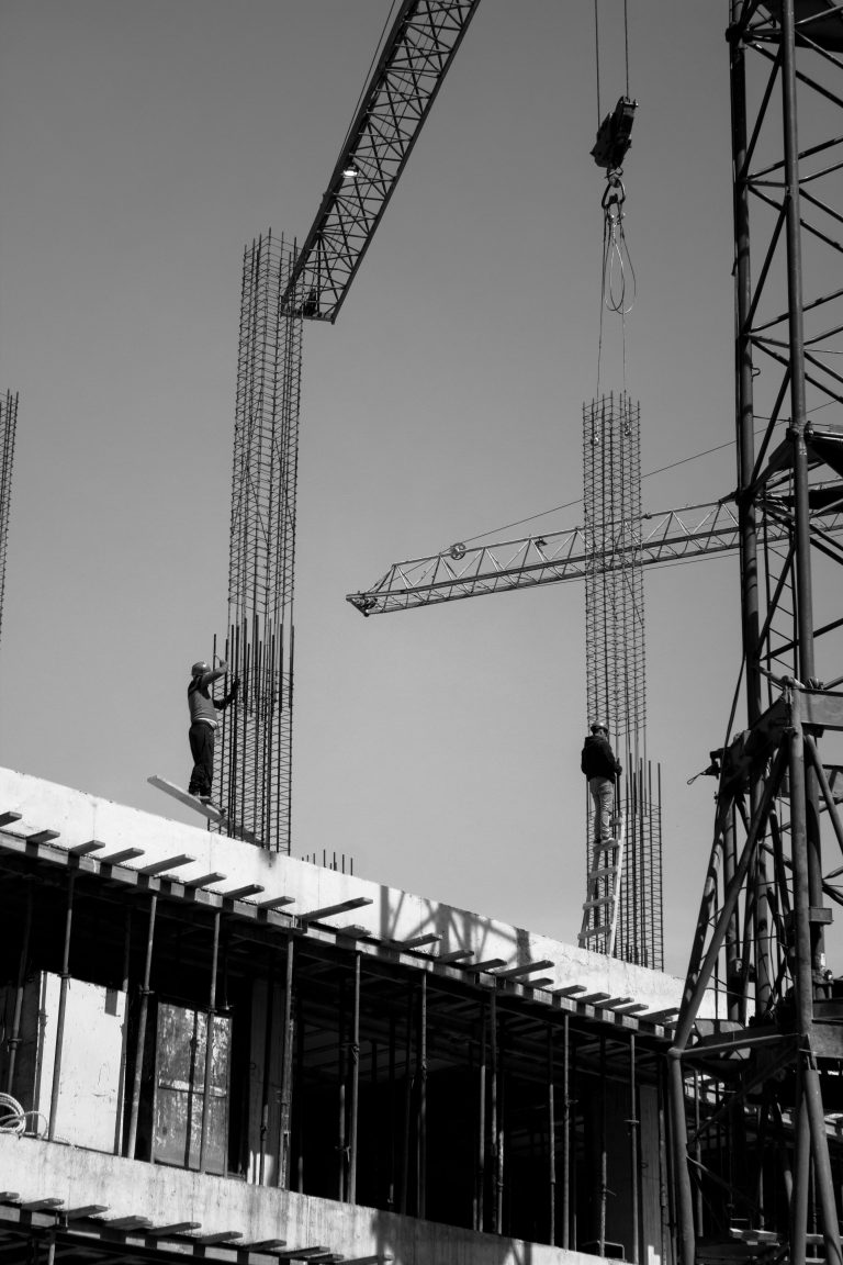 Black and white photo of two construction workers balancing on beams, with cranes and scaffolding rising against a clear sky. The scene conveys focus and precision.
