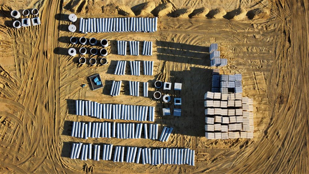 Aerial view of a construction site with neatly arranged stacks of concrete pipes and square blocks on sandy ground. Tire tracks form patterns, conveying organization.