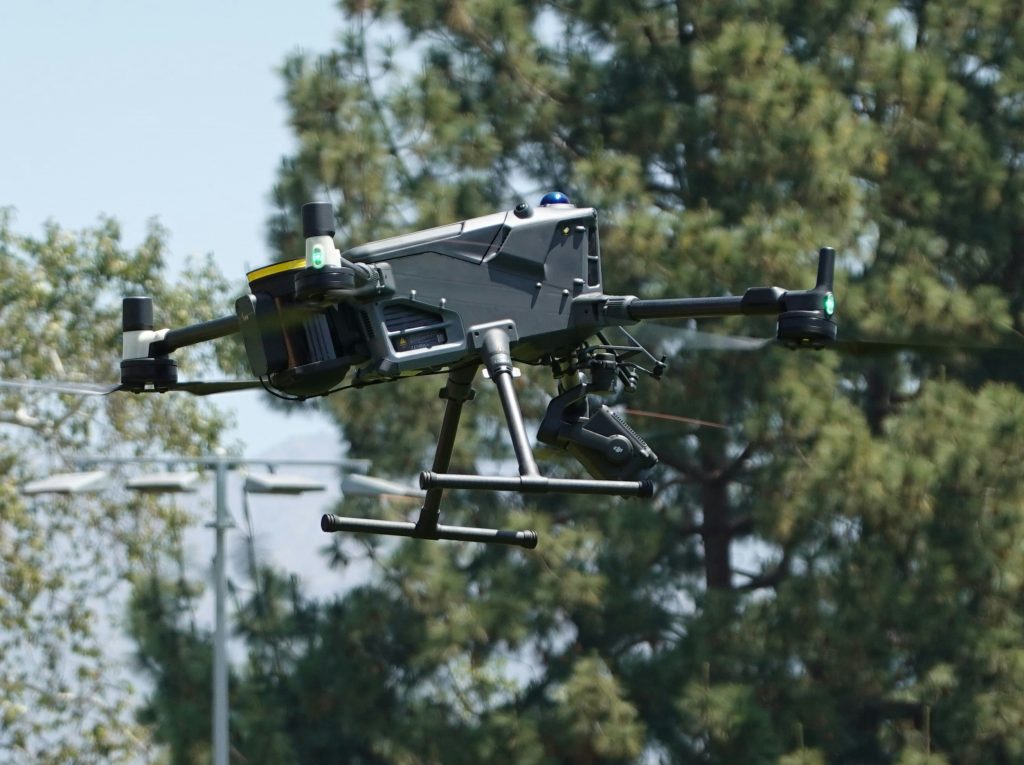 A black quadcopter drone with a mounted camera hovers mid-air against a backdrop of green trees and a clear sky, conveying a tone of modern technology in use.