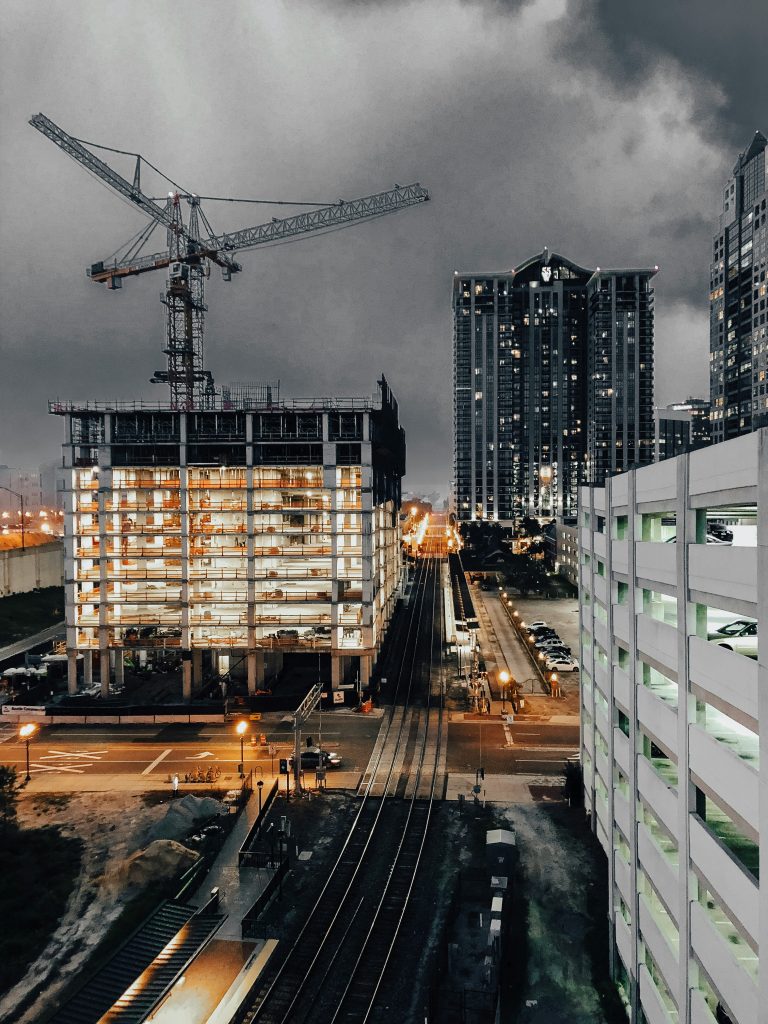 Construction site at dusk with a crane above a partially-built, illuminated building. Nearby are railway tracks and high-rise buildings under a cloudy sky.