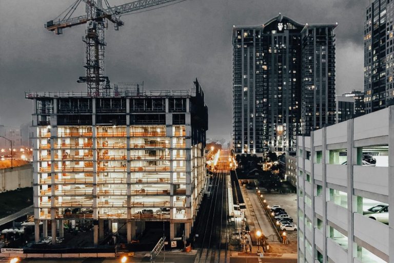Construction site at dusk with a crane above a partially-built, illuminated building. Nearby are railway tracks and high-rise buildings under a cloudy sky.