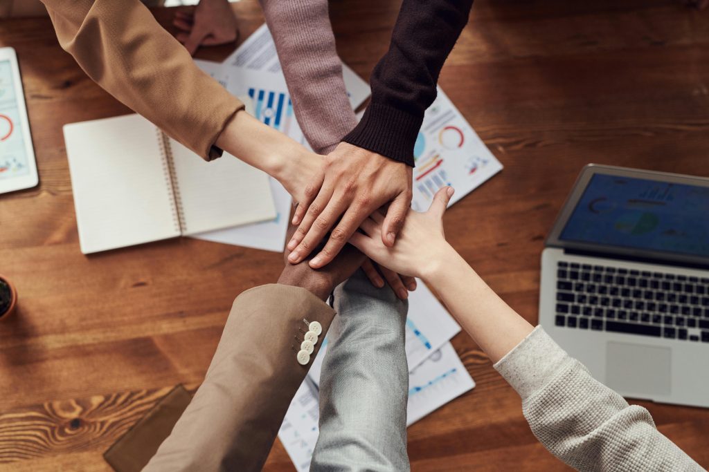 Hands of diverse individuals stacked in the center over a wooden table with documents and a laptop, symbolizing teamwork and collaboration.