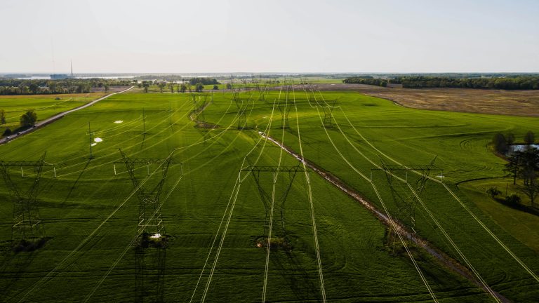 Aerial view of power lines stretching across a vast, green field under a clear sky. The lines create a network effect, conveying modern infrastructure in a rural landscape.