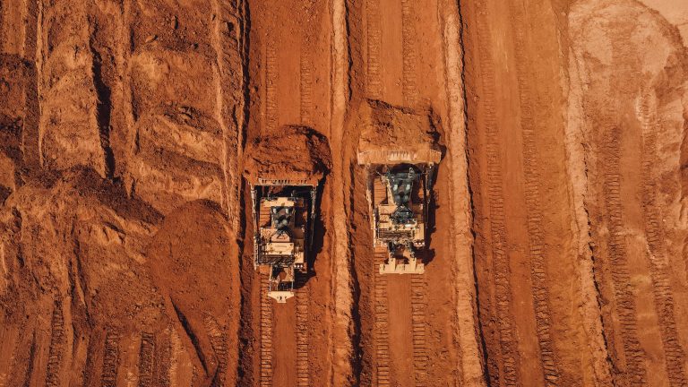 Two bulldozers move earth on a construction site. Aerial view shows parallel tracks in reddish-brown soil, conveying industry and progress.