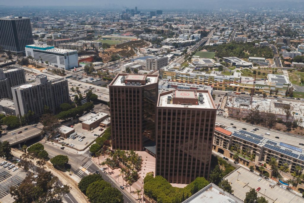 Aerial view of two tall, dark glass office buildings with helipads on top, surrounded by cityscape and roads under a hazy blue sky.