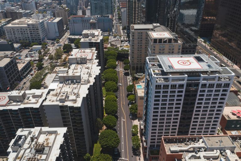 Aerial view of a cityscape with tall buildings featuring helipads. A road lined with green trees runs between the structures. Bright and urban setting.