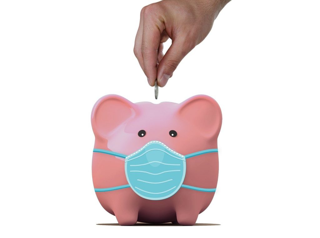 A pink piggy bank wearing a blue surgical mask is being used, as a hand above drops a coin into it, symbolizing saving money during uncertain times.